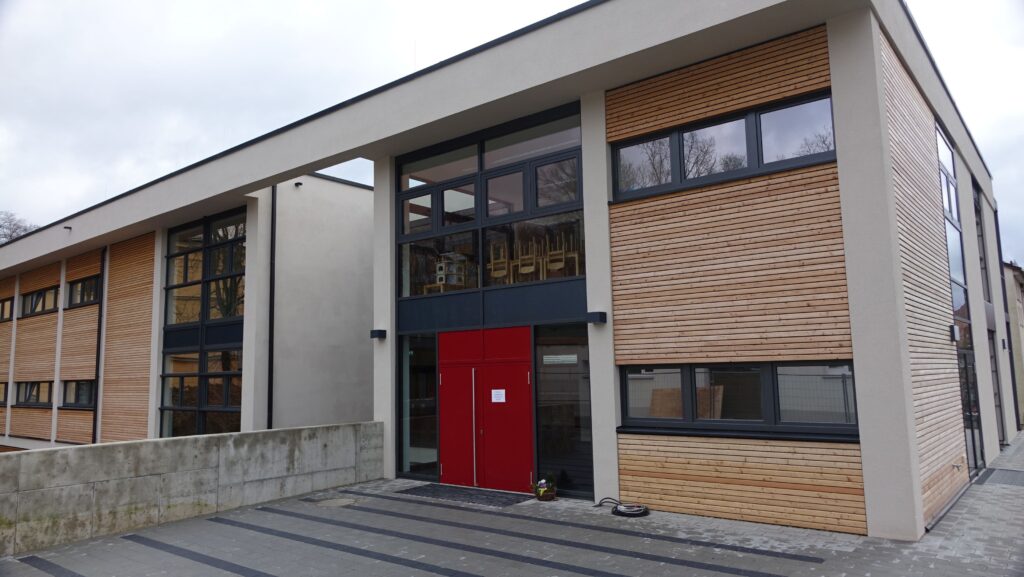 Eingangsbereich des Hortes "Grashüpfer": Die Eingangstür ist rot, die Fassade geprägt durch Glaselemente und Holzlamellen.