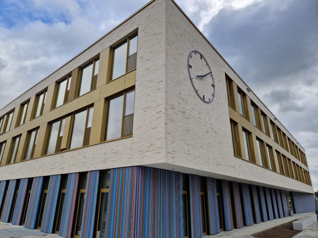 Hausansicht der neuen Grundschule "Magister-Hering": Grundschule und Hort teilen sich ein Gebäude. Die Fassade ist zweigeteilt: unten senkrechte bunte Metallstreben, oben Klinkerfassade mit integrierten Fensterband