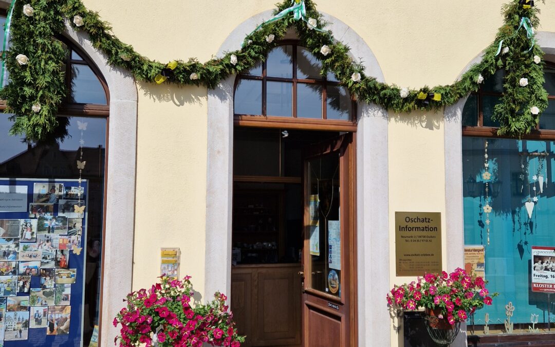Eingang zur Oschatz-Information mit Blumendekoration und Girlanden, das Schild über der Tür, sonniges Wetter, Fenster und Infotafeln sichtbar.