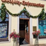 Eingang zur Oschatz-Information mit Blumendekoration und Girlanden, das Schild über der Tür, sonniges Wetter, Fenster und Infotafeln sichtbar.