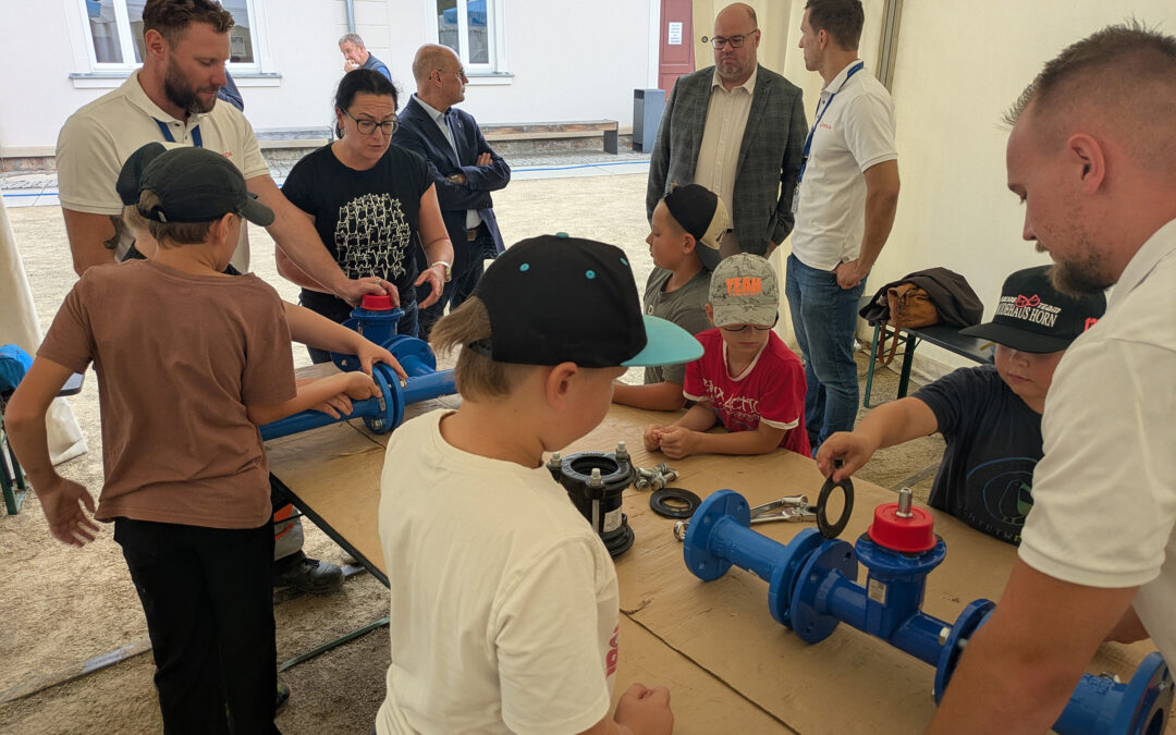 Kinder und Erwachsene arbeiten gemeinsam an blauen Rohrteilen und Armaturen unter einem weißen Zelt, um technische Fähigkeiten oder Wassertechnik zu entdecken.