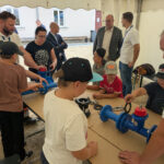 Kinder und Erwachsene arbeiten gemeinsam an blauen Rohrteilen und Armaturen unter einem weißen Zelt, um technische Fähigkeiten oder Wassertechnik zu entdecken.